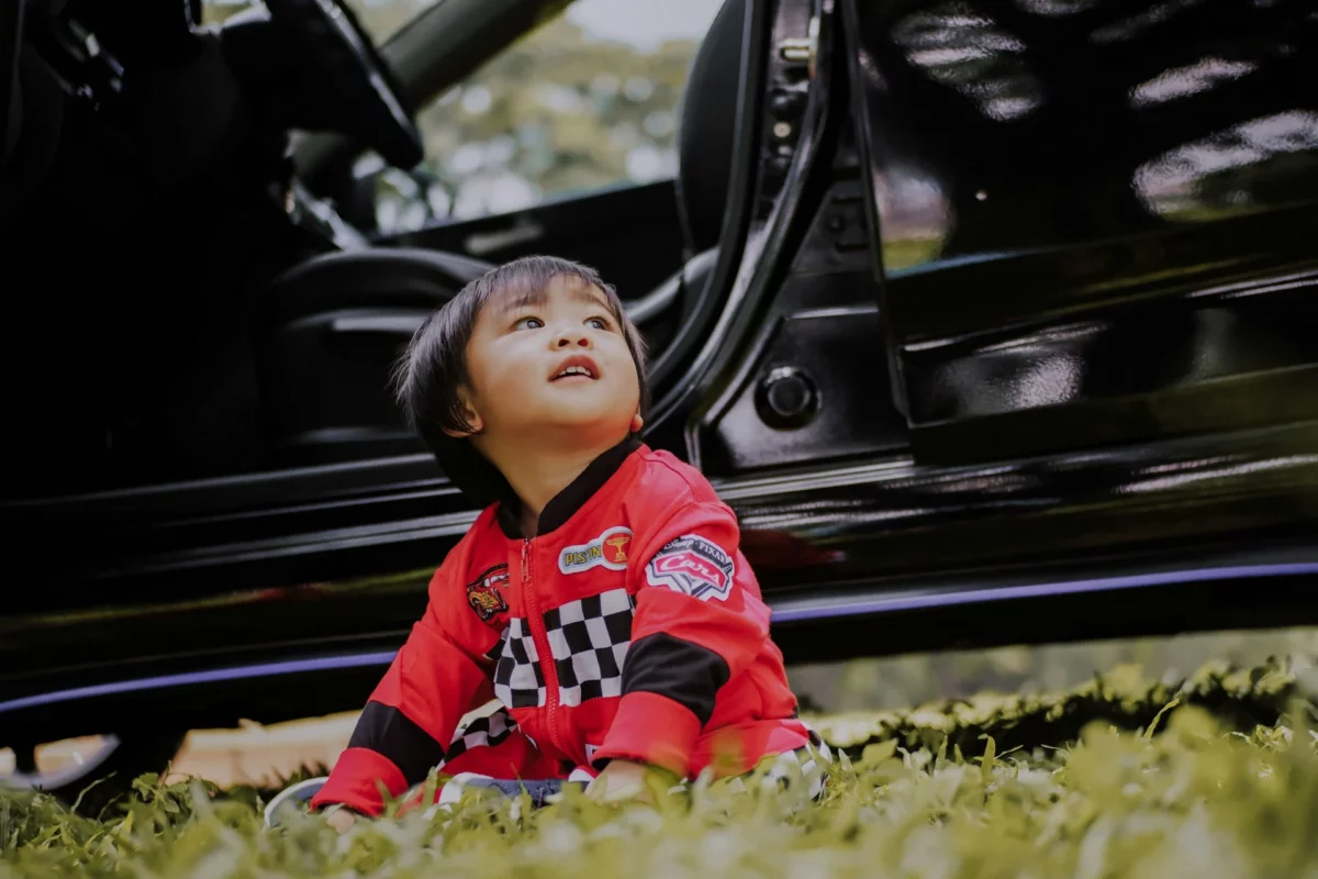 Toddler sitting near a parked car, dressed in a vibrant red jacket with a racing theme, highlighting the importance of child safety during car journeys.