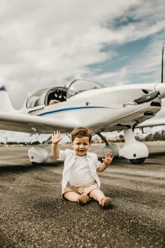 Smiling toddler sitting on the ground near an airplane, symbolizing stress-free family travel with compact gear like a cabin-sized stroller.