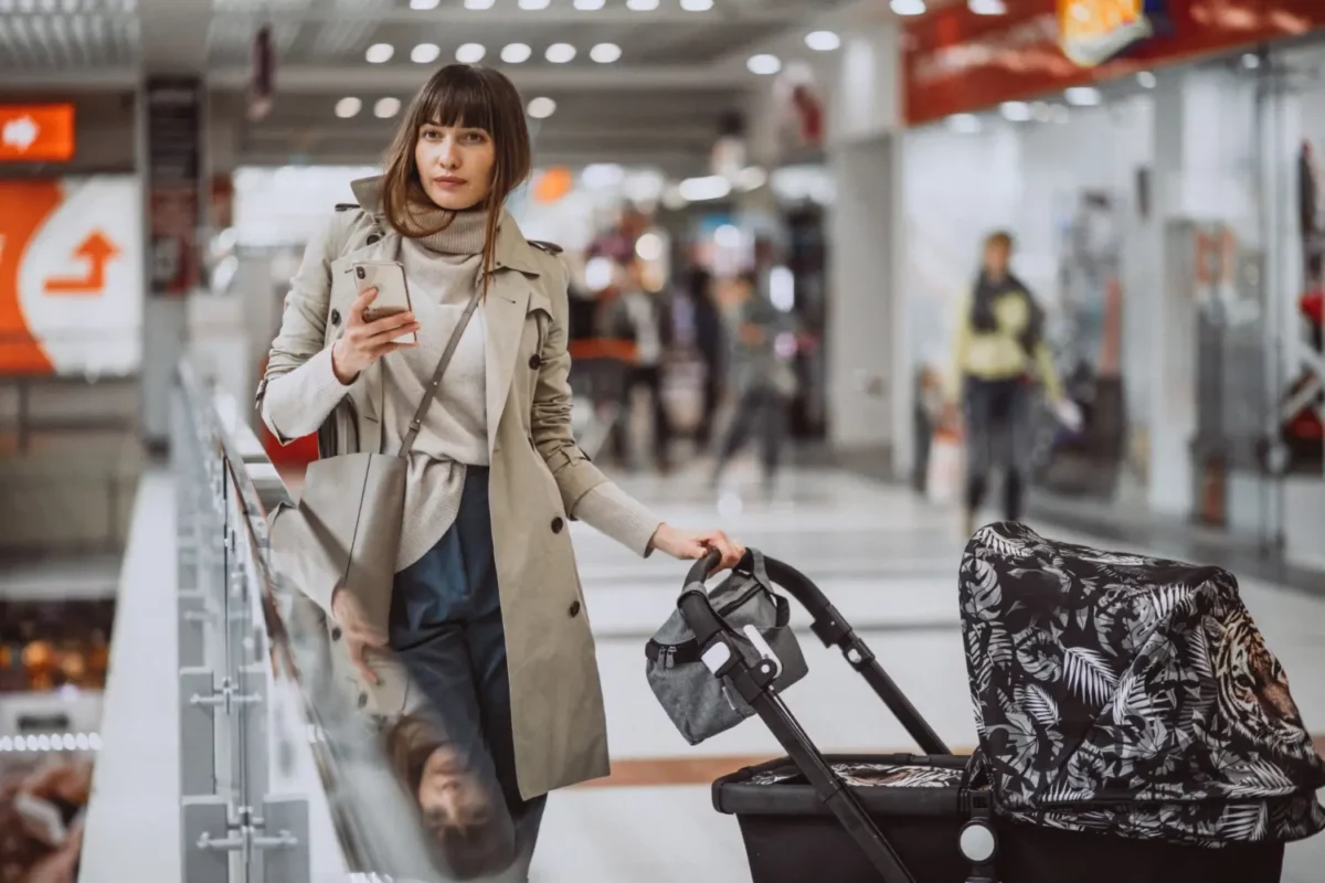 Stylish woman in an airport terminal holding her phone and pushing a designer stroller, showcasing modern travel convenience for parents.