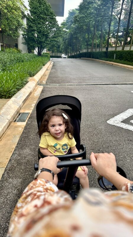 A happy toddler in a Koopers two-way stroller, parent-facing mode, enjoying a stroll in a lush outdoor setting.