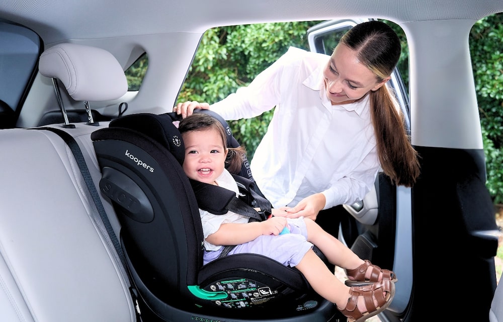 Happy toddler in a Koopers car seat with mother assisting inside a Malaysian car
