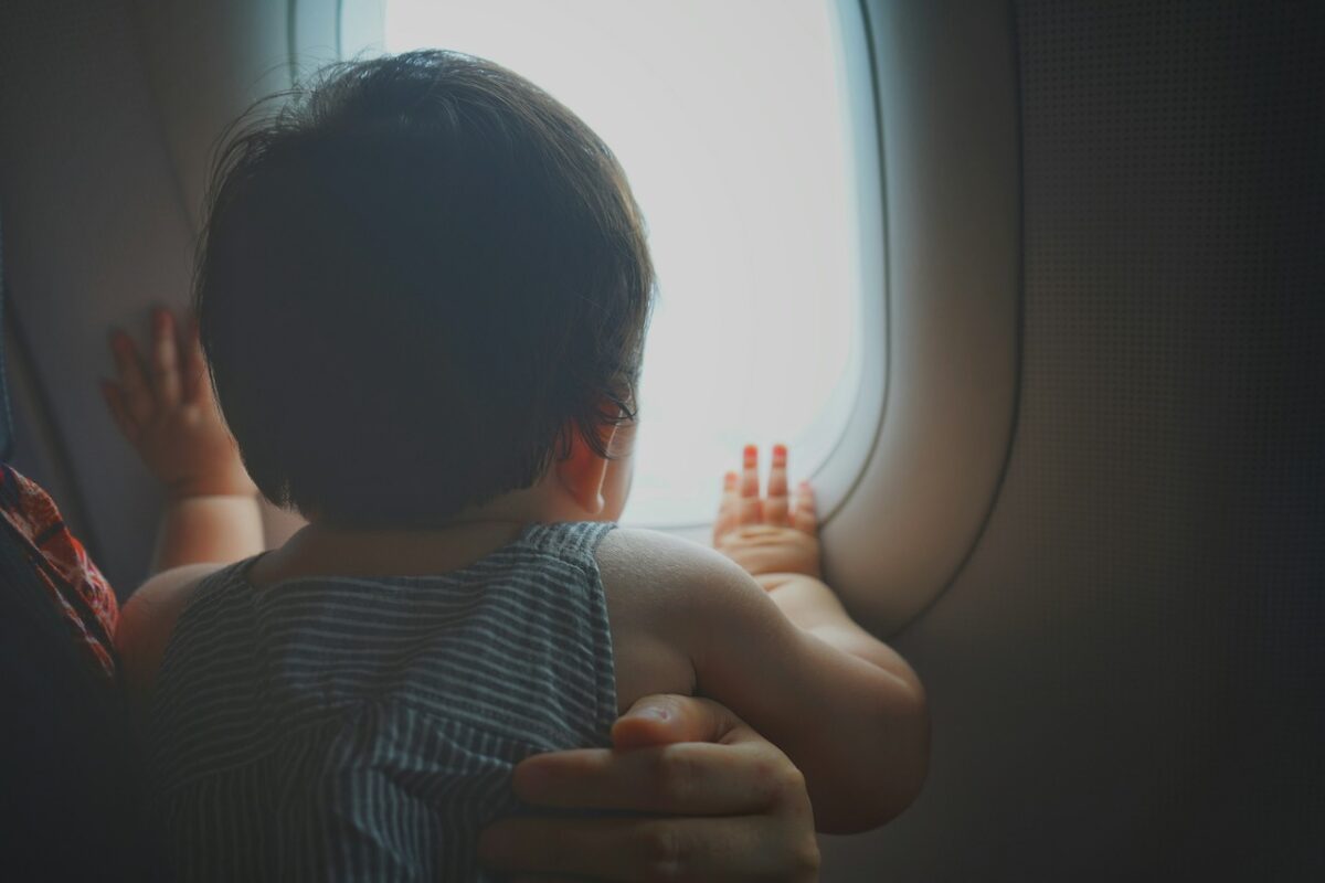 how-to-fly-with-a-car-seat-&-stroller-6 Baby in striped outfit gazing out of an airplane window while being held by an adult.