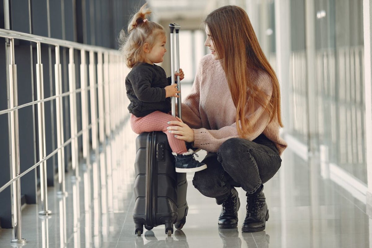Smiling toddler sitting on black suitcase while mother in pink sweater kneels beside her at the airport.