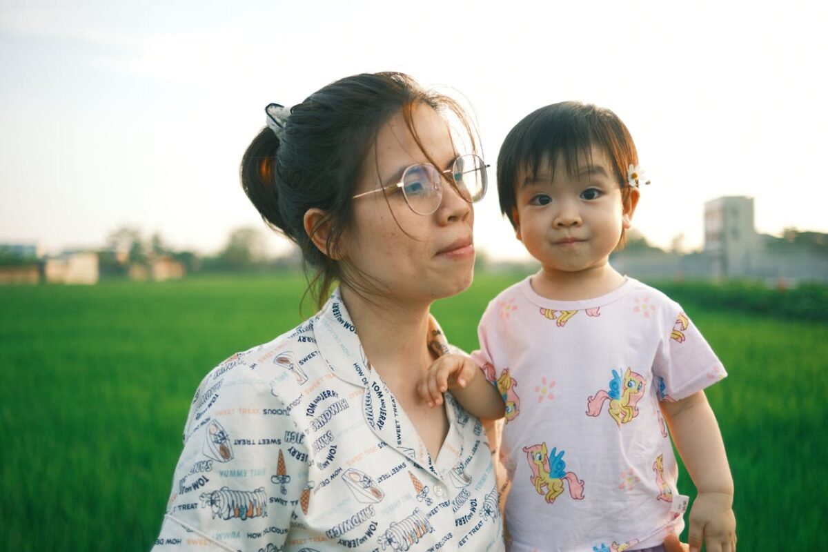 Mother and toddler standing in a green field during golden hour, both looking content and relaxed