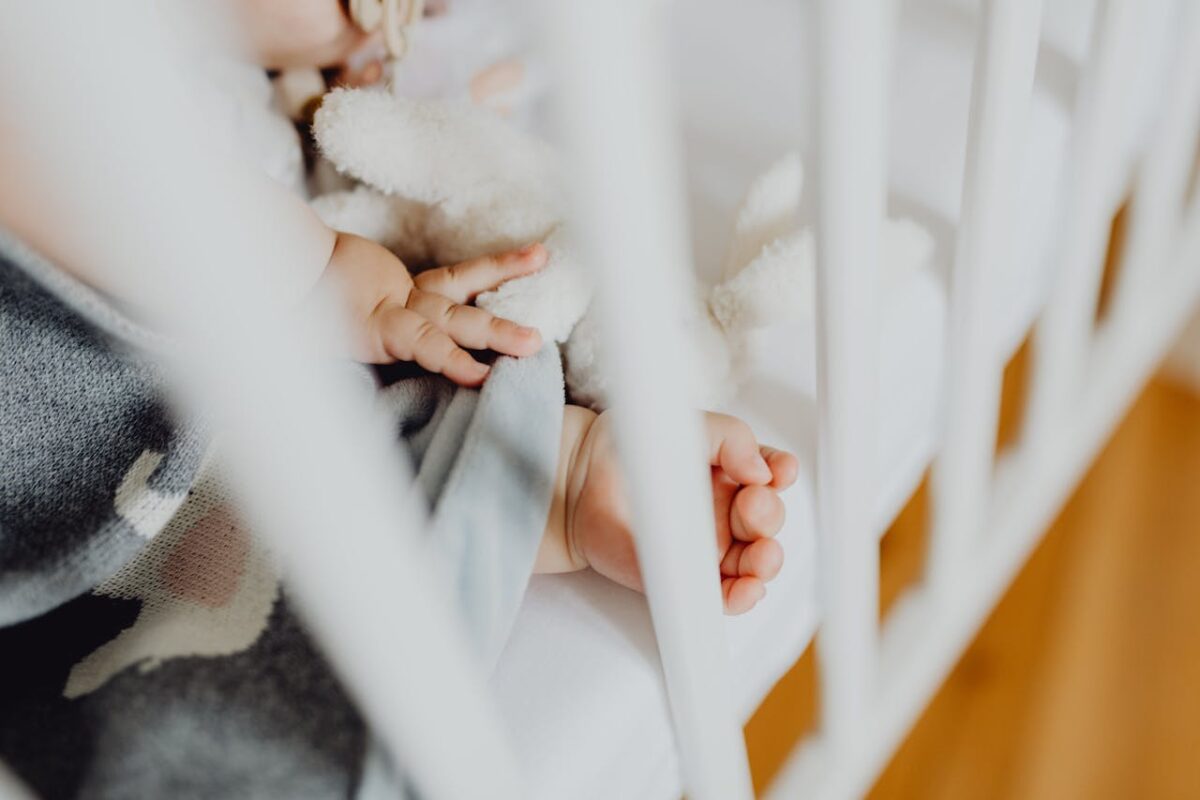 Close-up of baby’s hand gripping a soft toy inside a white baby cot, highlighting the importance of firm mattresses and safe sleeping setups over playpens for full-time sleep.