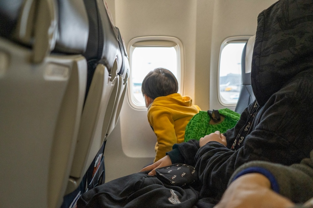 Child wearing yellow hoodie looking out airplane window during flight, family air travel with kids in Malaysia