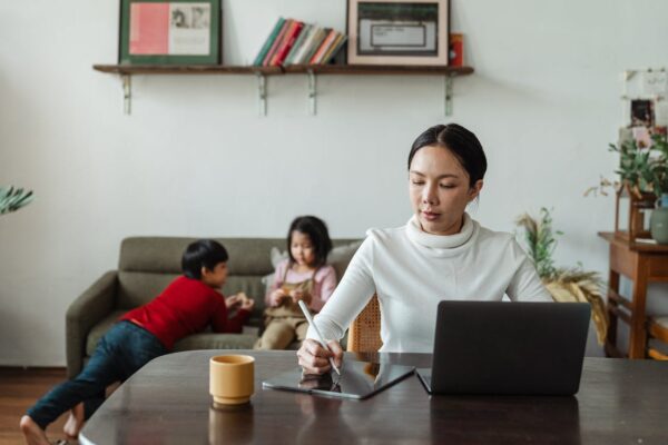 buying-baby-gear-online-vs-in-store-1 Malaysian mother researching baby gear on laptop at home with two young children playing nearby, representing online baby product shopping experience.