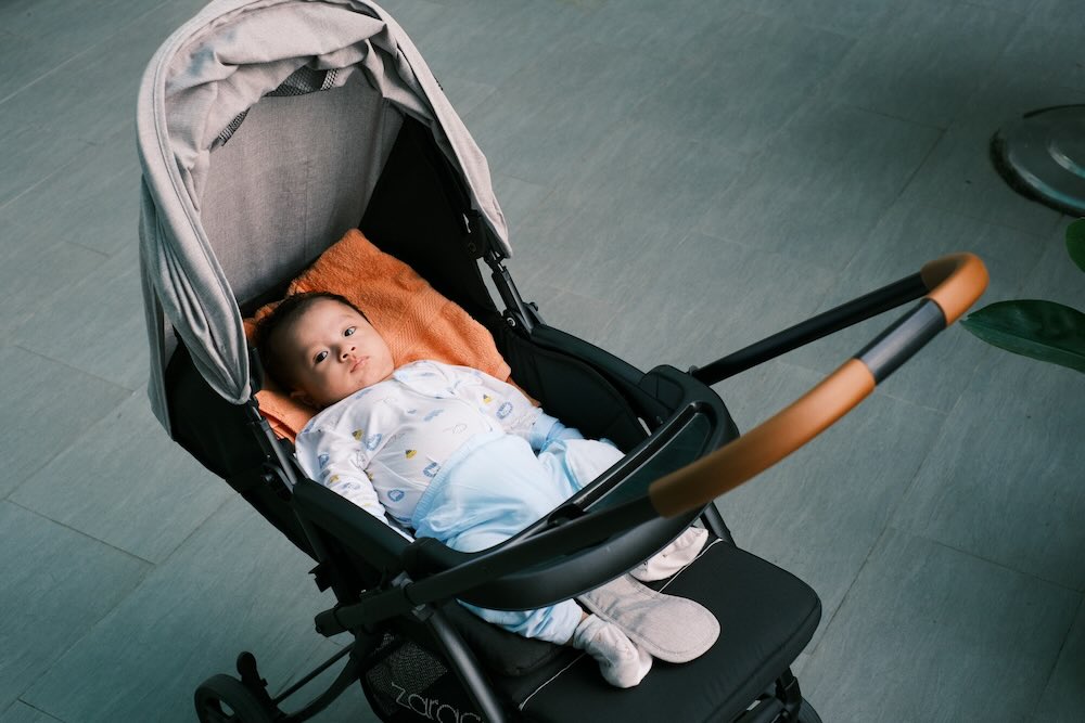 Baby lying in Koopers stroller with cushioned seat and canopy, ready for a stroll indoors or outdoors in Malaysia.