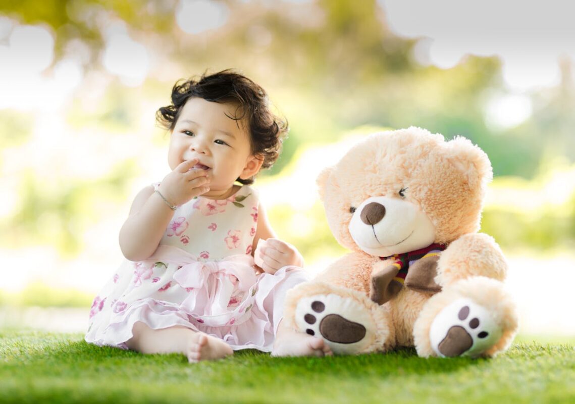 Smiling baby girl in a floral dress sitting on green grass beside a large teddy bear in a sunlit outdoor park setting.