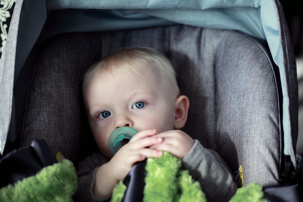 Baby with blue eyes in grey car seat holding green blanket, highlighting the need for secure and non-expired baby car seats.