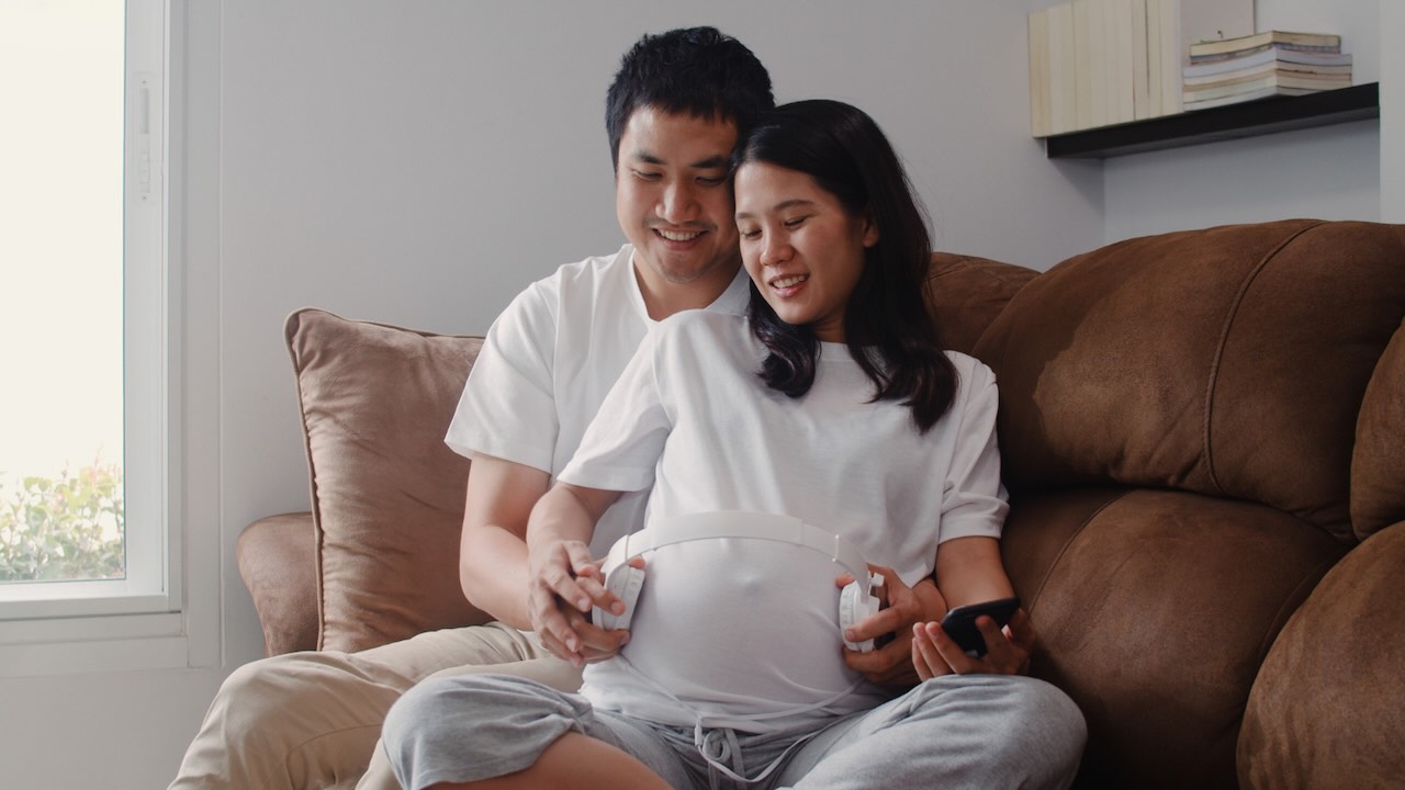 new-parents-tips-for-new-mums-dads-6 Expecting couple sitting together, smiling while placing headphones on the mother’s pregnant belly.