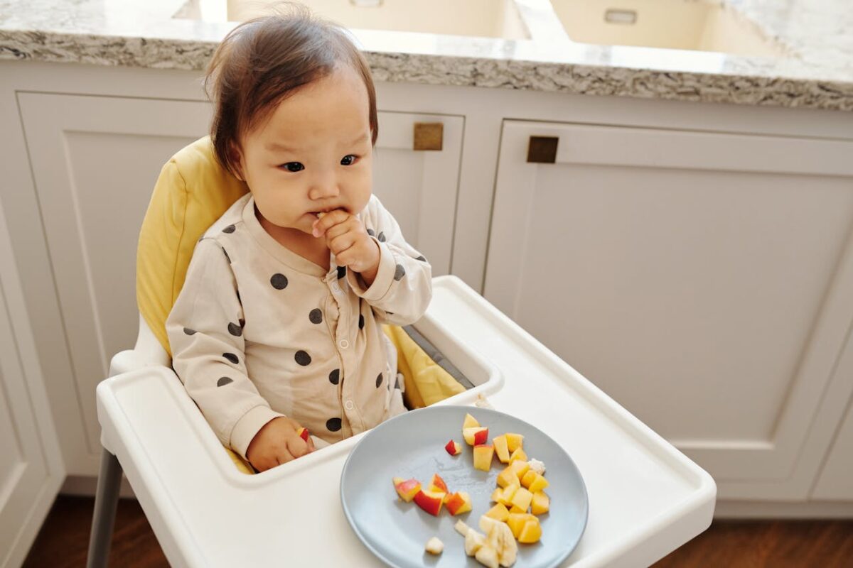Baby sitting in a high chair during mealtime, self-feeding soft fruit pieces in a bright kitchen setting.