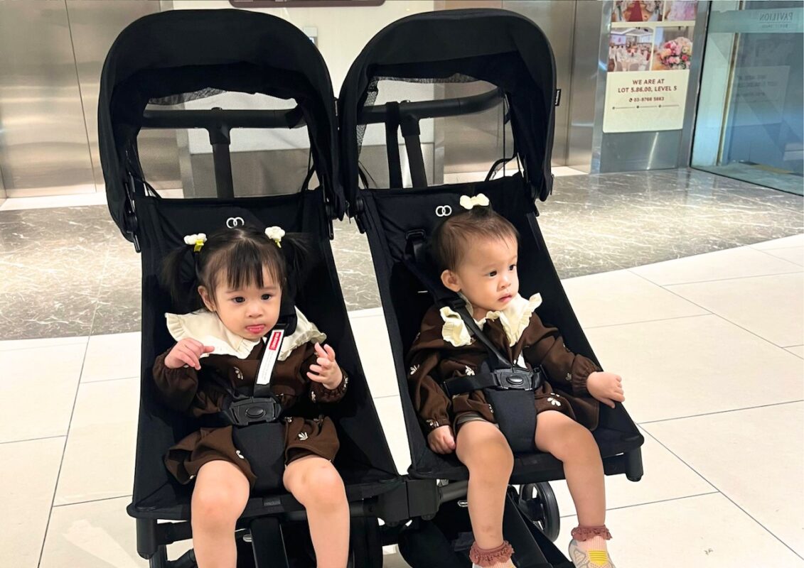 why-city-parents-need-smart-baby-stroller-car-seat-setup-1 Twin toddlers seated in side-by-side black strollers indoors, wearing matching outfits, looking around curiously in a mall setting