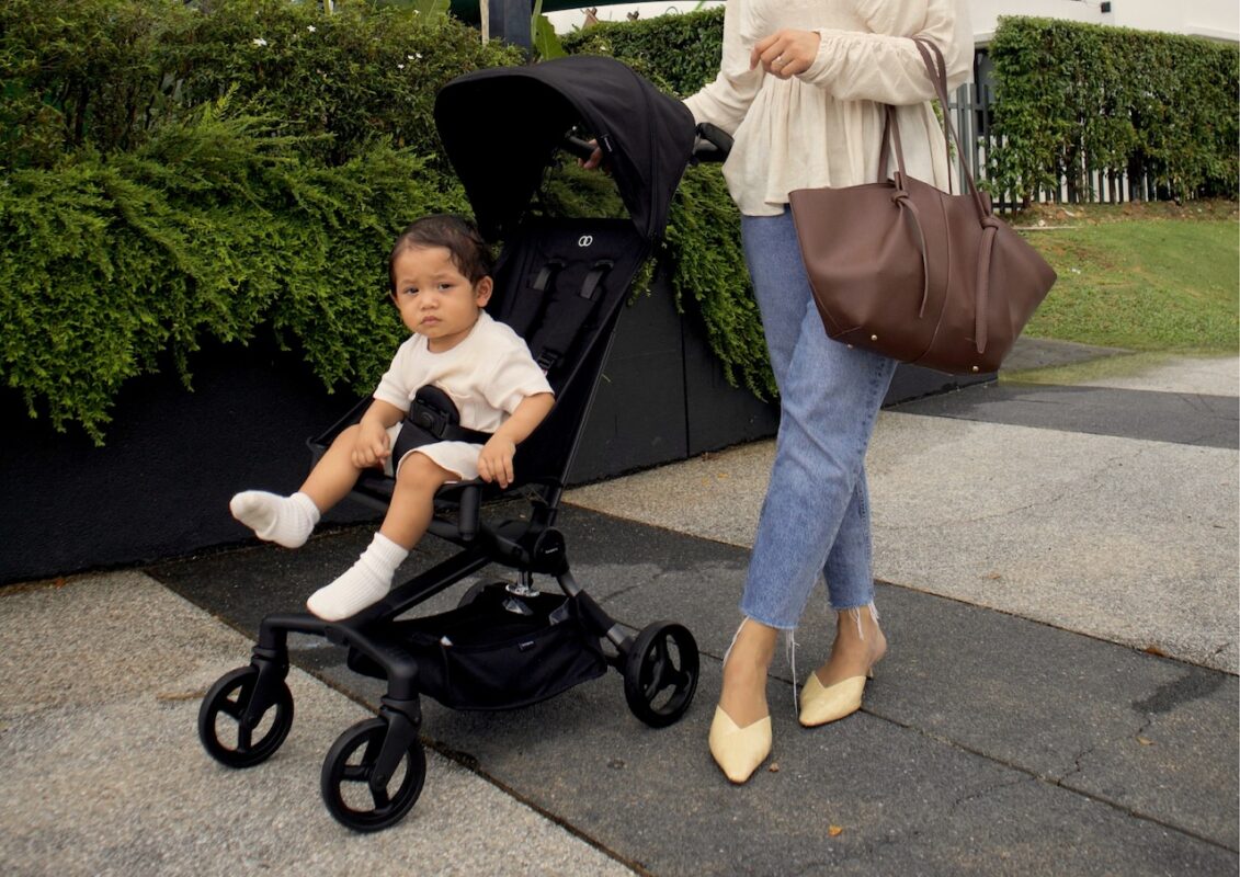 why-city-parents-need-smart-baby-stroller-car-seat-setup-6 Mother pushing toddler in black stroller along sidewalk, carrying brown handbag, with greenery and outdoor path in background