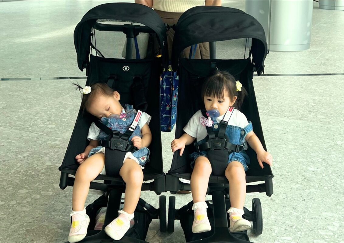 why-city-parents-need-smart-baby-stroller-car-seat-setup-7 Twin toddlers in matching outfits seated in side-by-side black strollers, one sleeping and one awake with a pacifier indoors
